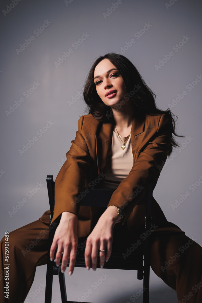 A pretty young woman with a smile on her face and wearing a brown classic suit is sitting on a chair on a gray background. Beautiful woman in a jacket and with makeup on her face