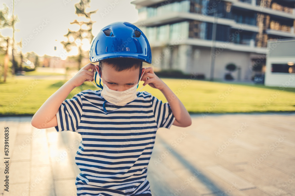 New Normal. Toddler boy on bicycle wearing protective mask and helmet. A boy adjusting a mask on his face while riding a bicycle. New Normal. Toddler boy on bicycle wearing protective mask and helmet. A boy adjusting a mask on his face while riding a bicycle.