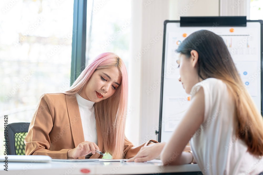 Two pretty young Asian businesswoman sitting at desk with laptop doing paperwork together discussing project financial report. Corporate business collaboration concept. Two pretty young Asian businesswoman sitting at desk with laptop doing paperwork together discussing project financial report. Corporate business collaboration concept.