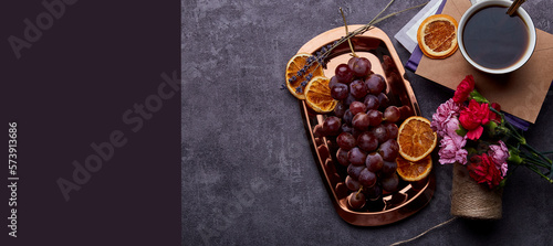 Aesthetic grapes on the golden tray with cup of coffee, dry oranges, books and flowers with copy space. Extra wide banner