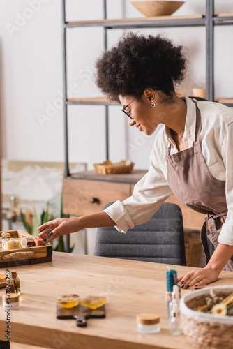 african american woman in eyeglasses and apron reaching soap bars near blurred ingredients on table in craft workshop.