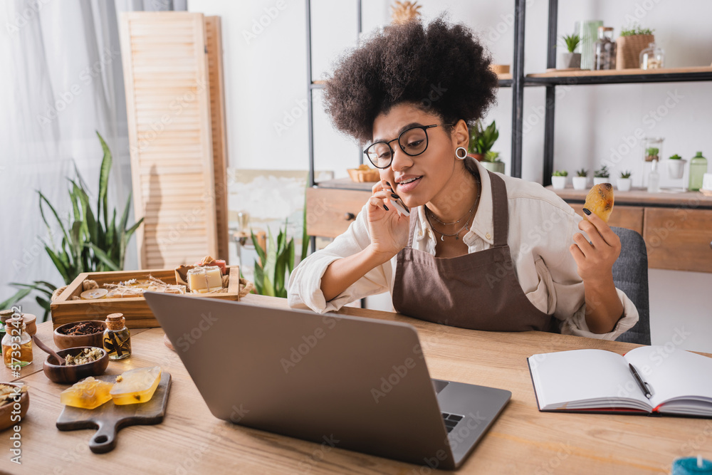 positive african american craftswoman talking on mobile phone near laptop and homemade soap in workshop. positive african american craftswoman talking on mobile phone near laptop and homemade soap in workshop.