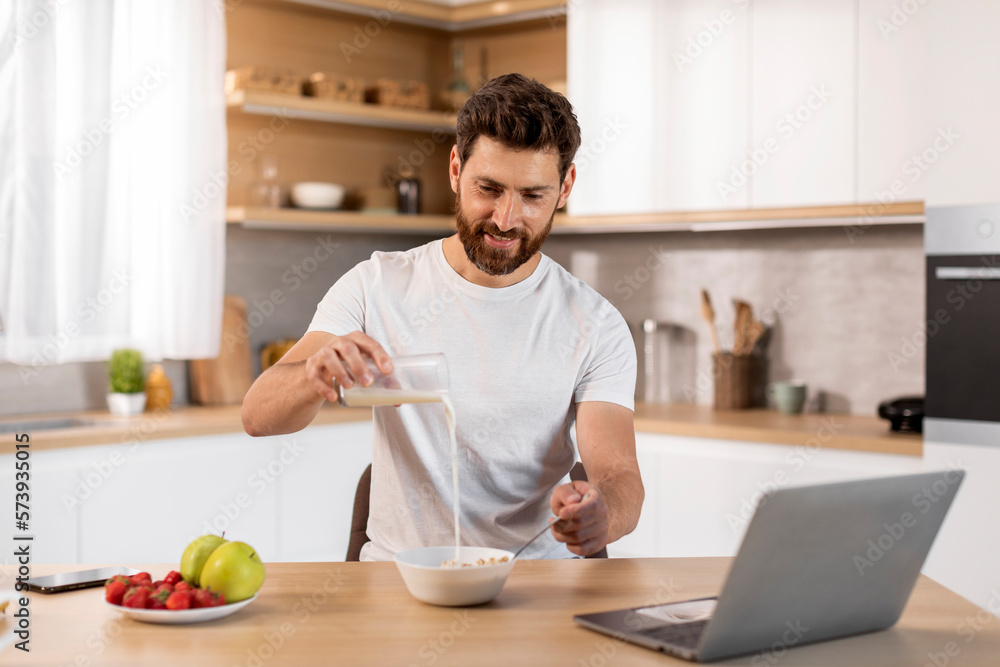 Smiling adult caucasian male in white t-shirt pours milk into cereal at table with laptop, watching video Smiling adult caucasian male in white t-shirt pours milk into cereal at table with laptop, watching video
