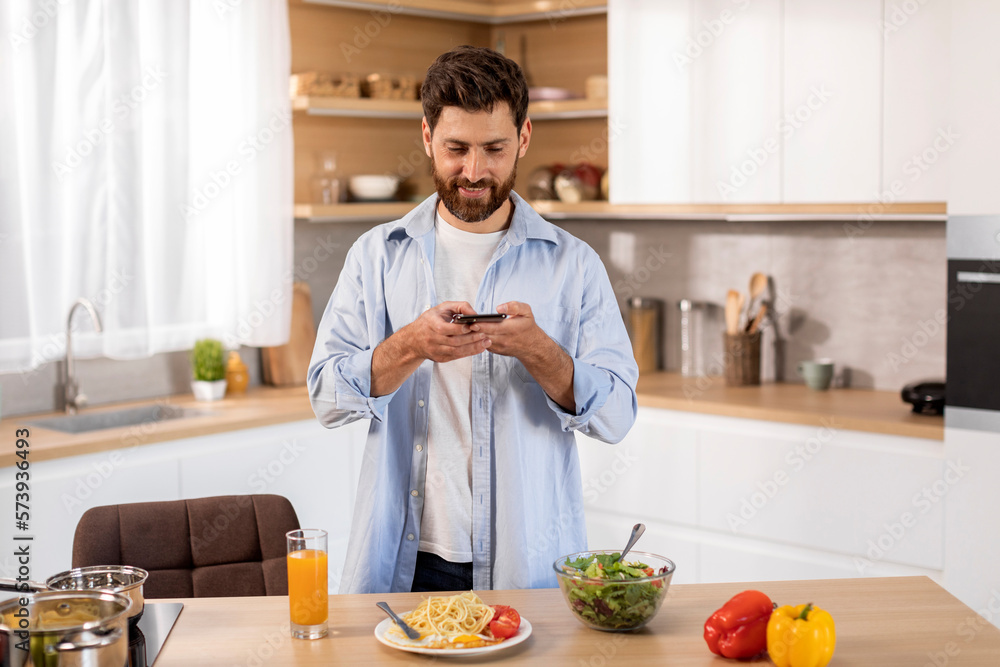 Smiling adult caucasian man in casual cooked dinner and takes pictures of pasta on smartphone Smiling adult caucasian man in casual cooked dinner and takes pictures of pasta on smartphone