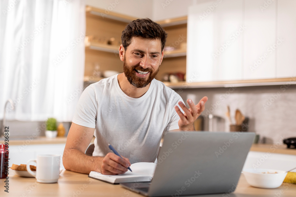 Happy busy adult caucasian male in white t-shirt has video call, making notes in modern kitchen interior Happy busy adult caucasian male in white t-shirt has video call, making notes in modern kitchen interior
