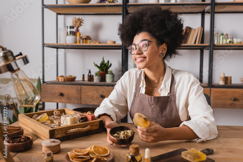 pleased african american craftswoman in eyeglasses holding bar of handmade soap and wooden bowl with dried herbs in workshop.