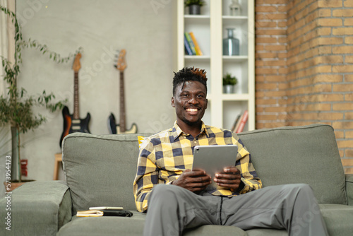 Young African man using digital tablet at home. Smiling man reading news while sitting on the couch at home photo