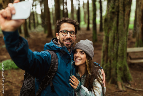 Cheerful millennial caucasian man hug lady in jacket and backpack, take selfie on phone in cold forest © Prostock-studio