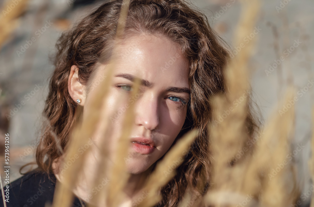 The face of a girl with dark curly hair is hidden behind dry grass. Blue eyes, black eyebrows. The face of a girl with dark curly hair is hidden behind dry grass. Blue eyes, black eyebrows.