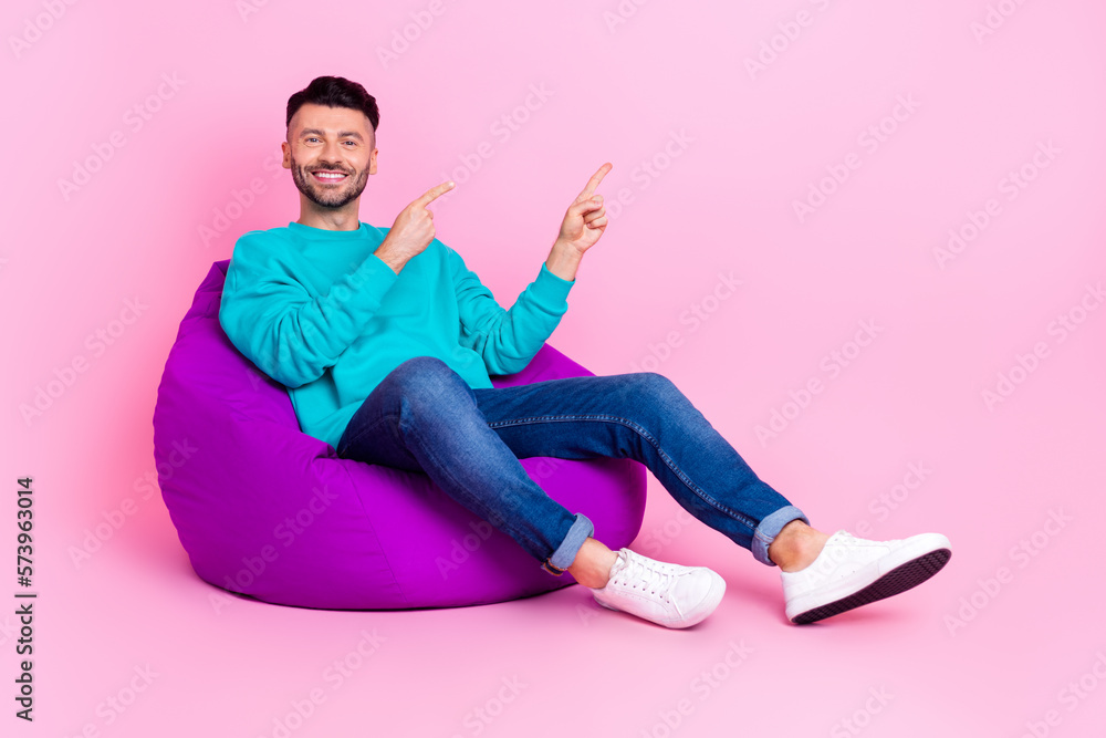 Full size photo of young handsome man sit soft bean bag point empty space dressed stylish blue clothes isolated on pink color background Full size photo of young handsome man sit soft bean bag point empty space dressed stylish blue clothes isolated on pink color background