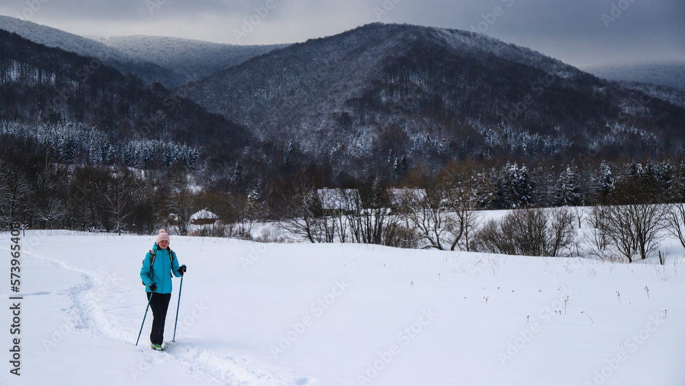 Hiker girl walks along snowy field with mighty mountains in background; hiking in cold winter weather