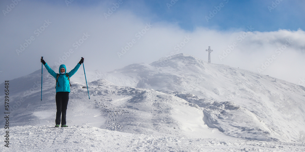A hiker girl with poles spreads her arms celebrating reaching the top of a mountain; extreme hiking in high mountains during the cold winter weather