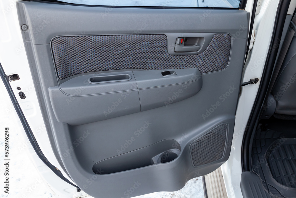 The open rear door of a Japanese car with black plastic trim and a gray textile insert with an automatic power window, a lock button on the panel and a storage compartment for items below.