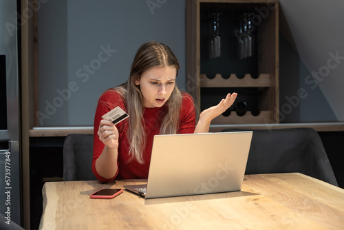 Young shocked frustrated woman holding bank credit card annoyed that she has reached her account limit and has no more money, she is refused online shopping and ordering on laptop computer