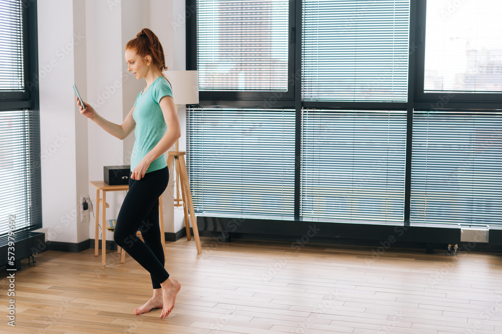 Fototapeta premium Wide shot of pretty sporty female vlogger in sportwear making video call using mobile phone on background of window at home. Redhead young woman exercising and making video call with personal trainer.