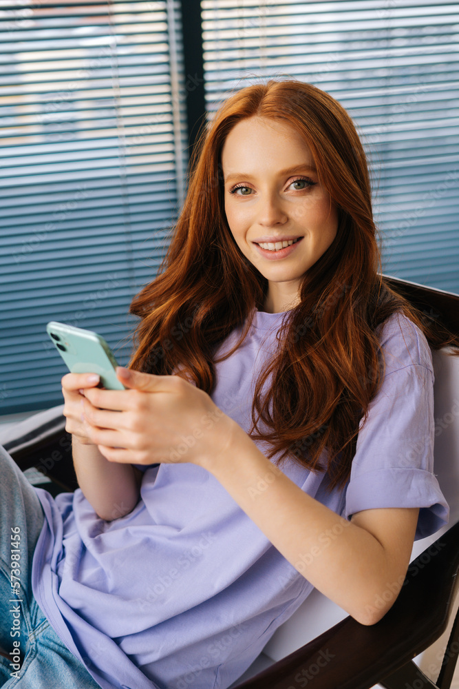 Vertical shot of pretty young woman texting online message using smartphone sitting on comfy armchair and smiling looking at camera. Cheerful female using cellphone at home on background of window. Vertical shot of pretty young woman texting online message using smartphone sitting on comfy armchair and smiling looking at camera. Cheerful female using cellphone at home on background of window.