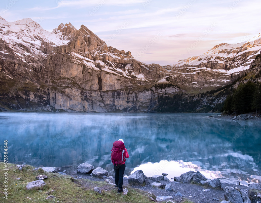 A hiker girl with backpack near mountain lake in Switzerland on sunrise with cloudy mountain peaks in the background. Lake Oeschinensee in Swiss alps