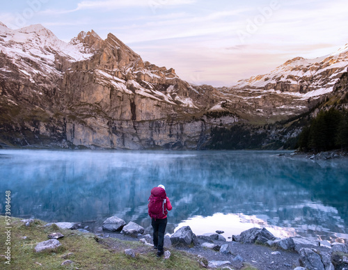 A hiker girl with backpack near mountain lake in Switzerland on sunrise with cloudy mountain peaks in the background. Lake Oeschinensee in Swiss alps