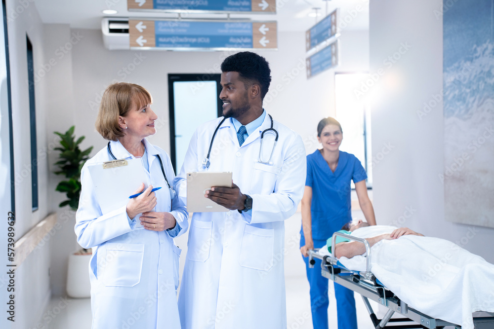 Obraz premium Selective focus of smiling mid-adult Caucasian female and young African male doctors, walking discussing over a tablet on hospital aisle with a blurred smiling nurse push a patient's bed in background