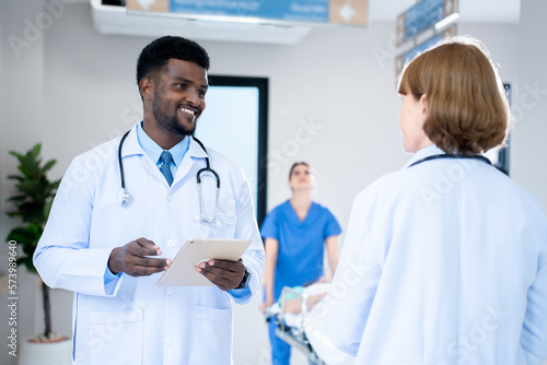 Selective focus of a smiling young African male medicine doctor in a white coat  standing holdilng a tablet and talking to a female doctor on the hospital corridor with a blurred nurse in background.