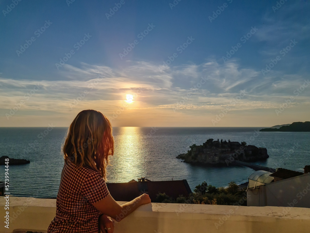 Happy woman on balcony in luxury hotel in Budva Riviera with scenic view on idyllic Sveti Stefan island at sunset, Adriatic Mediterranean Sea, Montenegro, Europe. Summer vacation in seaside resort