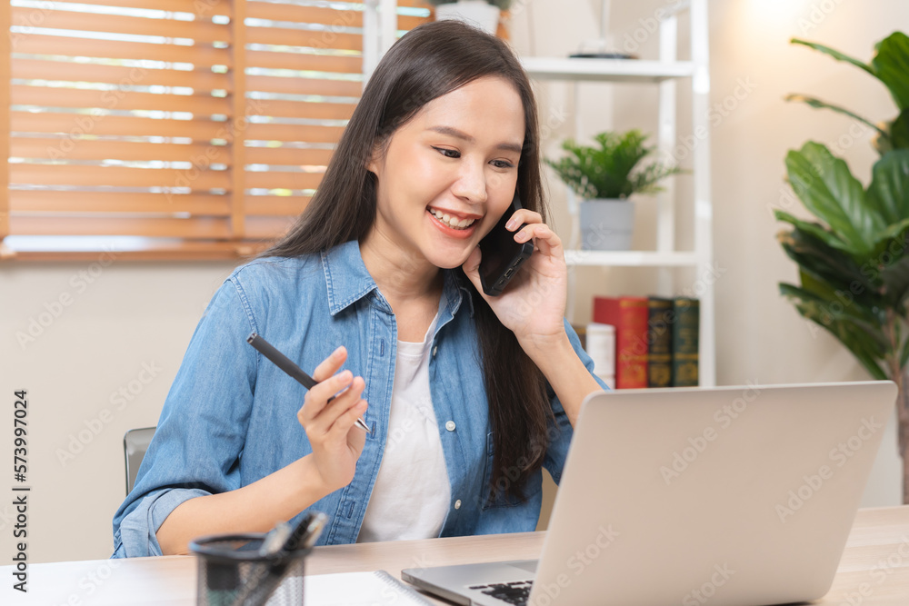 Smiling, entrepreneur asian young professional business woman discussing on call mobile, using device laptop computer on desk, holding smartphone talking to client while sitting work at home office. Smiling, entrepreneur asian young professional business woman discussing on call mobile, using device laptop computer on desk, holding smartphone talking to client while sitting work at home office.