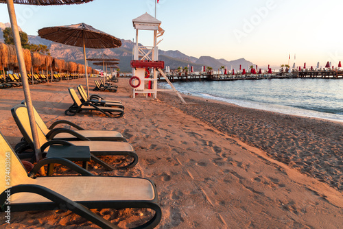 Fototapeta Naklejka Na Ścianę i Meble -  a row of lounge chairs sitting on top of a sandy beach, grand majestic mountains, turkey, wood pier and houses, sundown, stand up with the sea behind, sunbathing at the beach on Antalya Turkey	