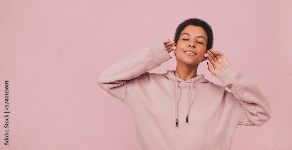 Fototapeta premium Stylish young african woman in hoodie lstening to music in wireless headphones. Lifestyle concept with copy space. Girl standing with closed eyes and enjoying songs over pink pastel backdrop 