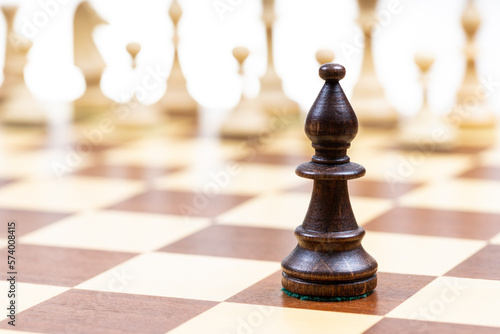 Tablou canvas black bishop against white chess pieces in background on wooden chessboard close