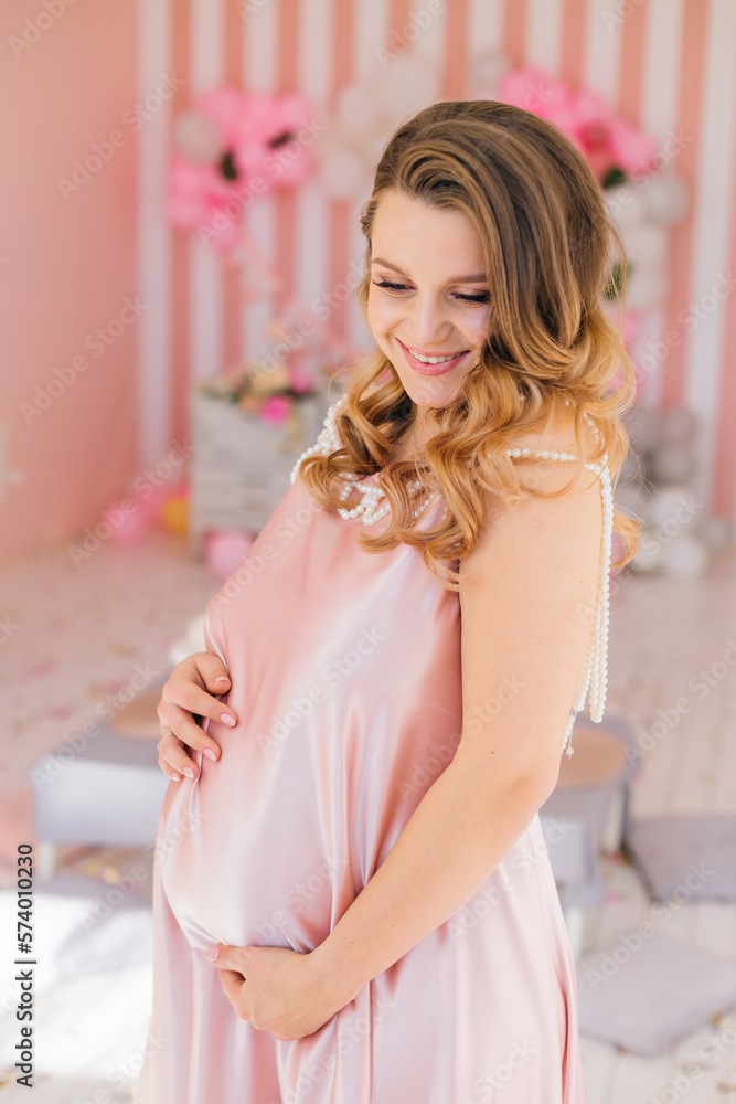 Naklejka premium A beautiful pregnant woman in a light pink dress is smiling and posing on a pink background of a children's room, waiting for a baby.