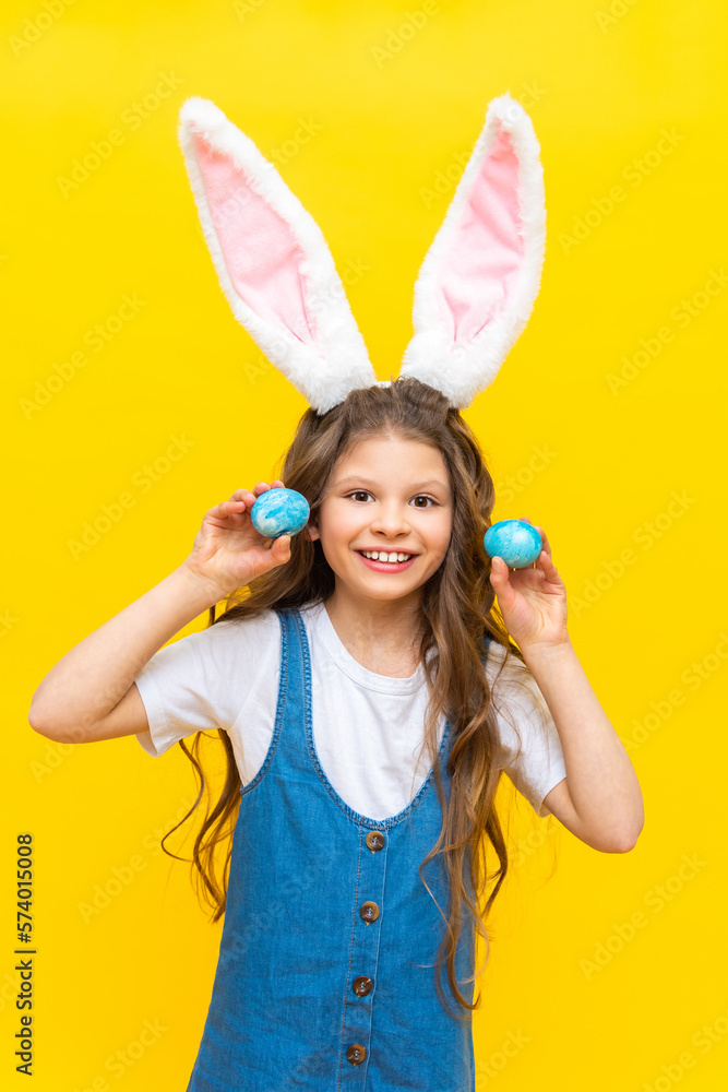 Happy Easter. A little girl holds two painted eggs for the holiday. A charming baby with rabbit ears on a yellow isolated background. Happy Easter. A little girl holds two painted eggs for the holiday. A charming baby with rabbit ears on a yellow isolated background.