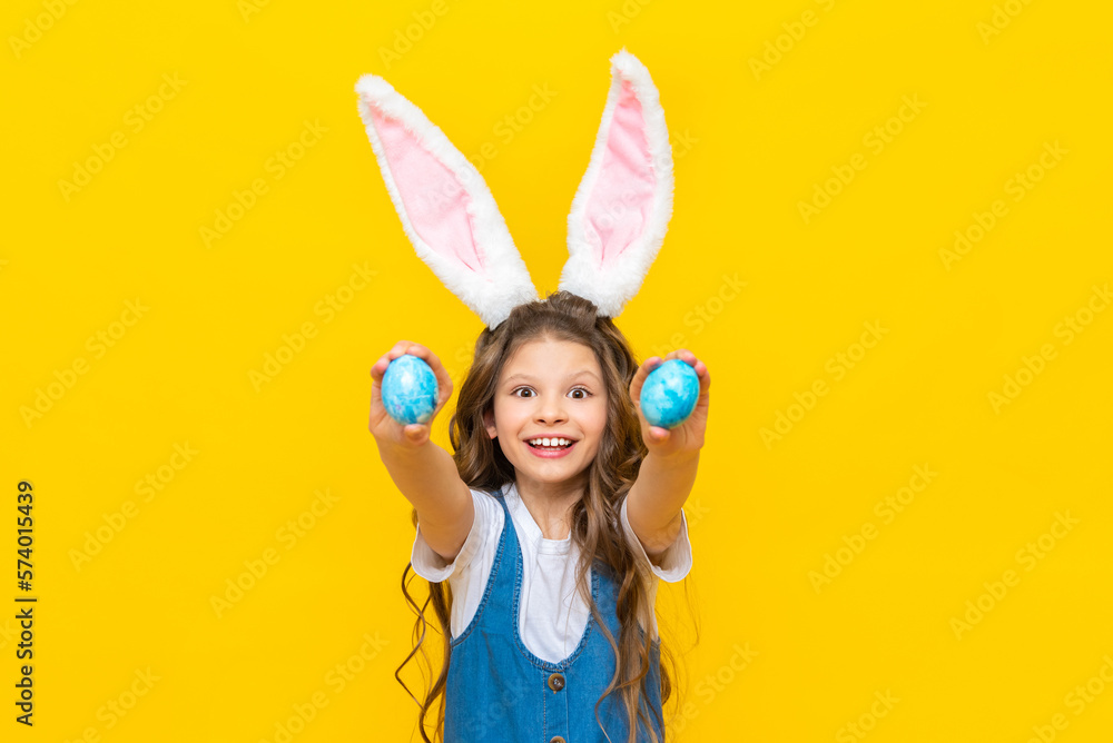 Fototapeta premium Happy spring Easter. A little girl with long hair holds two eggs and stretches them forward, smiling broadly. A charming child with rabbit ears in a blue sundress on a yellow isolated background.