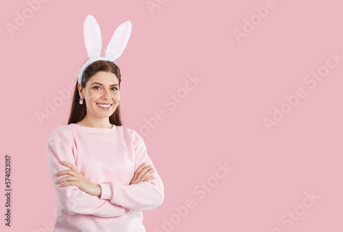 Happy Easter bunny girl on pink copy space background. Beautiful young woman wearing pastel pink sweatshirt, earrings and funny rabbit ears standing with her arms folded on pink copy space background
