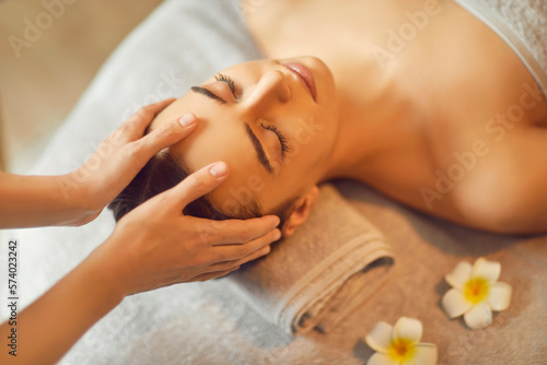 Beautiful young woman receiving facial massage in spa salon. Beautiful girl lying on massage bed on her back with closed eyes enjoying massage. Facial beauty treatment, body care, pampering