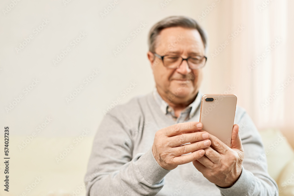 People And Technology. Close up smiling mature man using mobile phone, in living room at home. Elderly man using mobile phone. Focus on hand. People And Technology. Close up smiling mature man using mobile phone, in living room at home. Elderly man using mobile phone. Focus on hand.