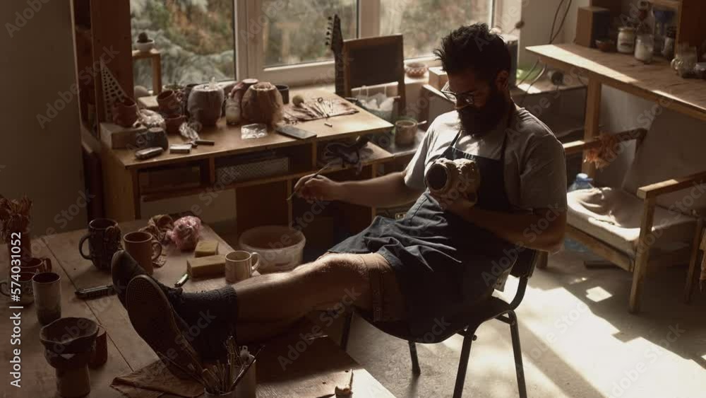 Bearded confident focused man in glasses making a jug from clay in his own pottery workshop, needlework, making handmade tableware. Bearded confident focused man in glasses making a jug from clay in his own pottery workshop, needlework, making handmade tableware.