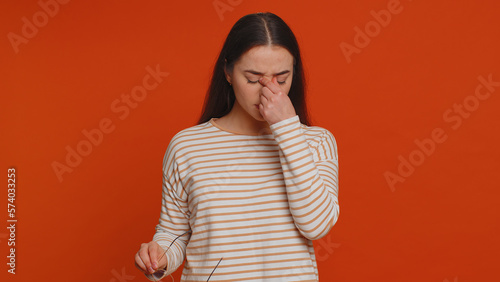 Exhausted tired pretty woman takes off glasses, feels eyes hard pain, being overwork burnout from long hours working, headache. Unhappy sad young adult girl isolated alone on red studio background