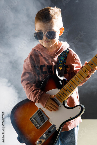 Chłopiec z gitarą - dziecko 5 lat gra na gitarze elektrycznej - mały rockman w okularach słonecznych w studiu z dymem