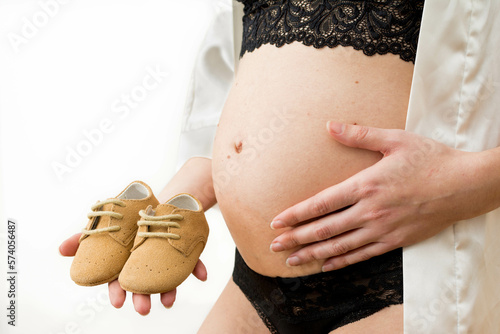 Tiny Steps to Parenthood: Unrecognizable Pregnant Woman Holding Baby Shoes in White Studio Background