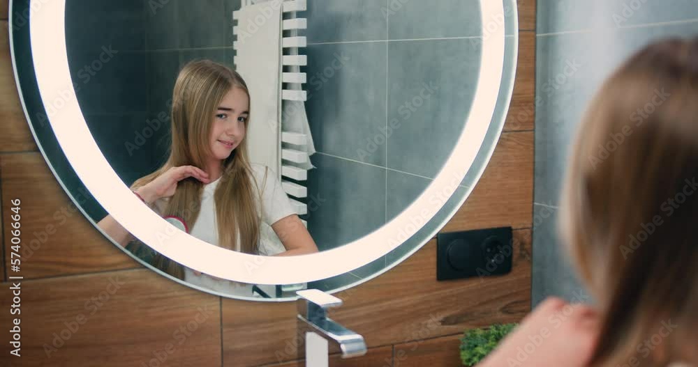 Little attractive girl brushing her hair and assembles in a pigtail in the bathroom. The girl looks directly into the mirror. Little attractive girl brushing her hair and assembles in a pigtail in the bathroom. The girl looks directly into the mirror.