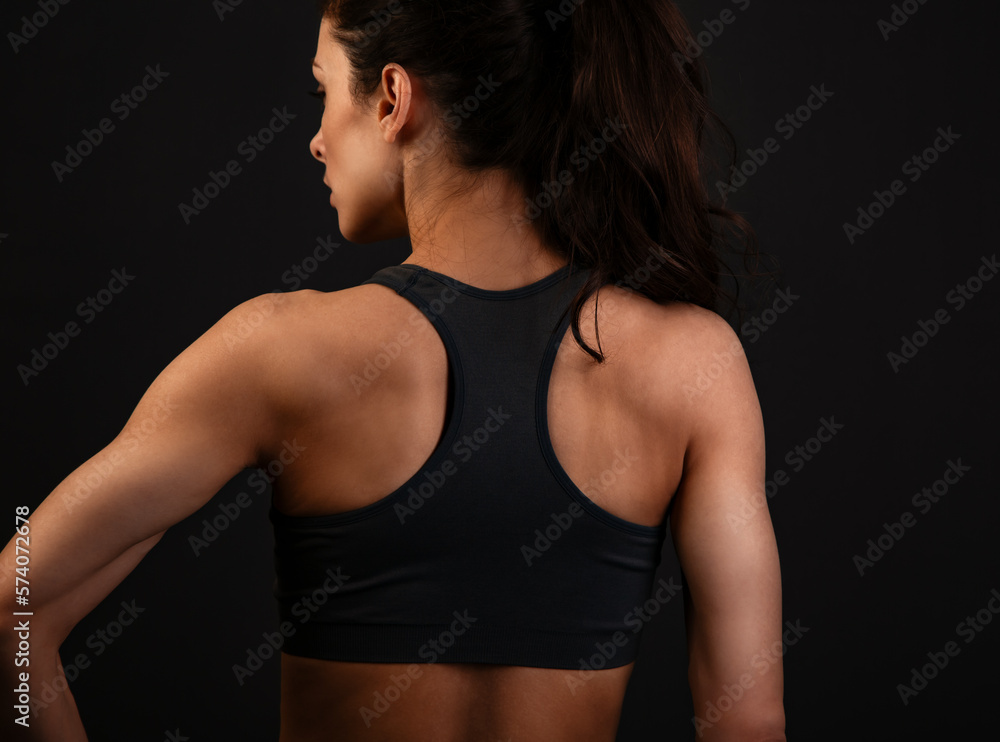 Serious female sporty muscular with ponytail doing stretching workout the shoulders, blades and arms in sport bra, standing on dark grey background with empty copy space. Back view. Serious female sporty muscular with ponytail doing stretching workout the shoulders, blades and arms in sport bra, standing on dark grey background with empty copy space. Back view.