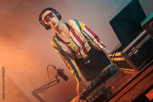 Flirty woman DJ acting silly with tongue out, fooling around in studio with lights. Playful funky musician mixing techno music with mixer or turntables, being confident and laid back. photo