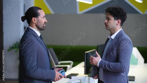 Two man employees in suits are talking in the office, a boss and a worker, solving work tasks. A man is holding a laptop.
