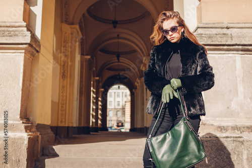 Stylish woman wearing fur coat with green gloves holding handbag on background of city architecture. Spring fashion.