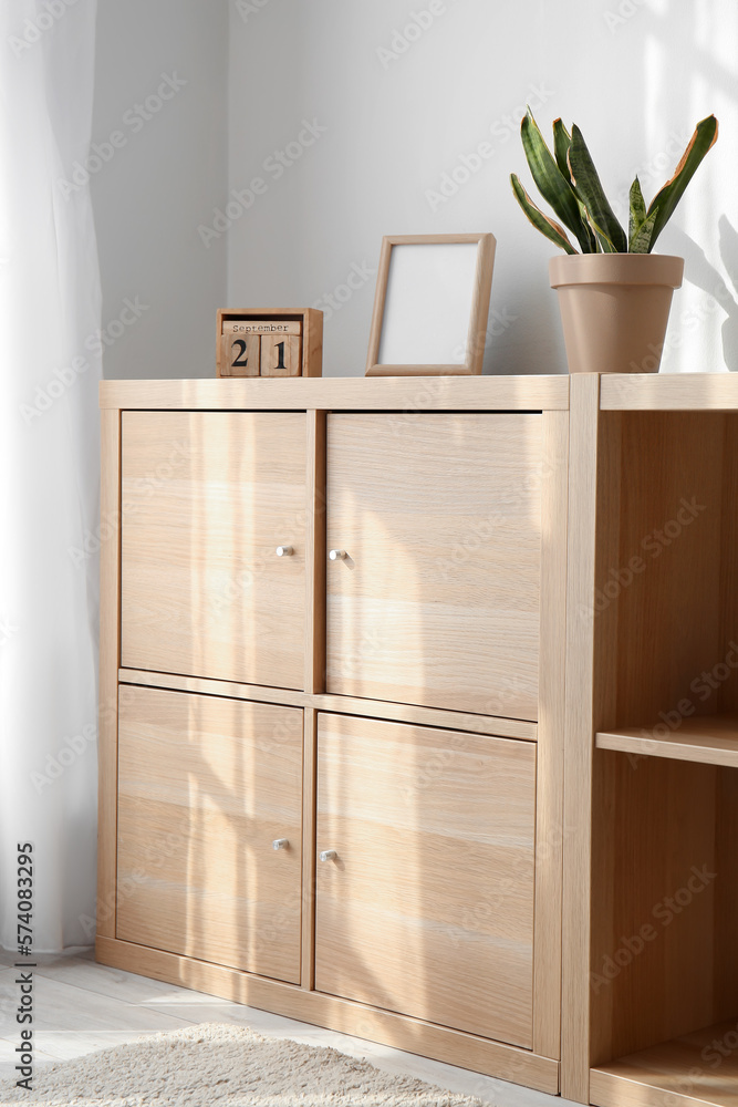 Fototapeta premium Wooden chest of drawers with calendar, frame and houseplant near light wall