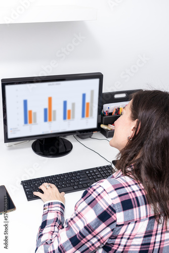 Close-up rear view of young business woman working in office interior on pc on desk, typing, looking at screen with diagrams