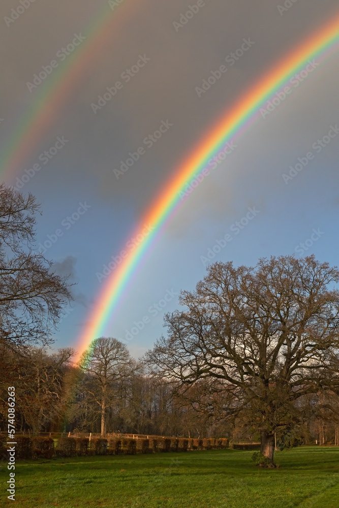 Himmel mit dunklen Wolken und doppeltem Regenbogen mit intensiven Farben und einem angedeuteten überzähligen Bogen innerhalb des Hauptregenbogens über dem Jersbeker Park in Schleswig-Holstein.