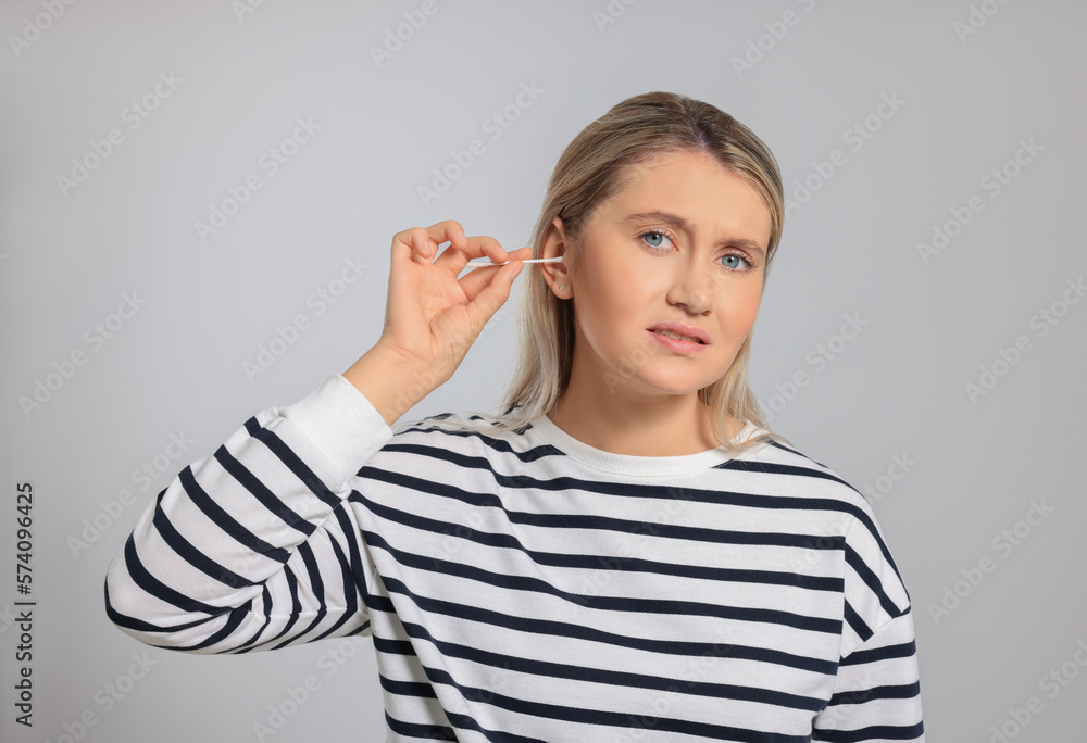 Young woman cleaning ear with cotton swab on light grey background Young woman cleaning ear with cotton swab on light grey background