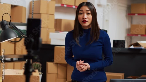 Young entrepreneur filming teleshopping video on phone, advertising products in warehouse space. Female worker using social media channel to create online ad promotion. Handheld shot.
