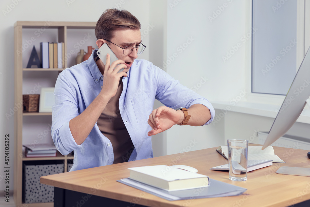 Emotional young man checking time while talking on phone in office. Being late Emotional young man checking time while talking on phone in office. Being late